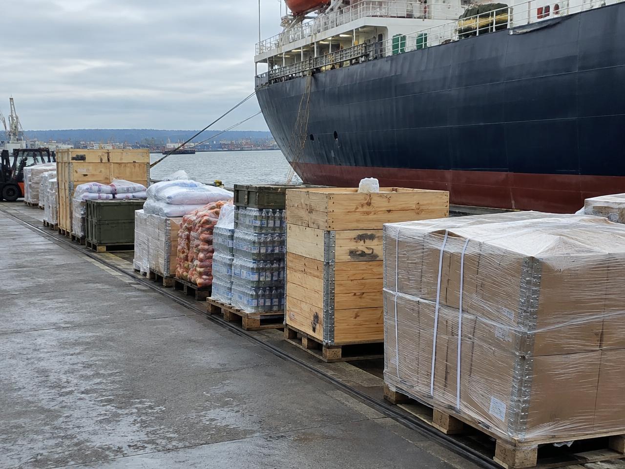 Illustrative photo: palletised ship supplies beside a merchant vessel at a commercial port